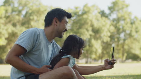 Happy-father-sitting-on-grass-with-daughter-and-taking-selfie