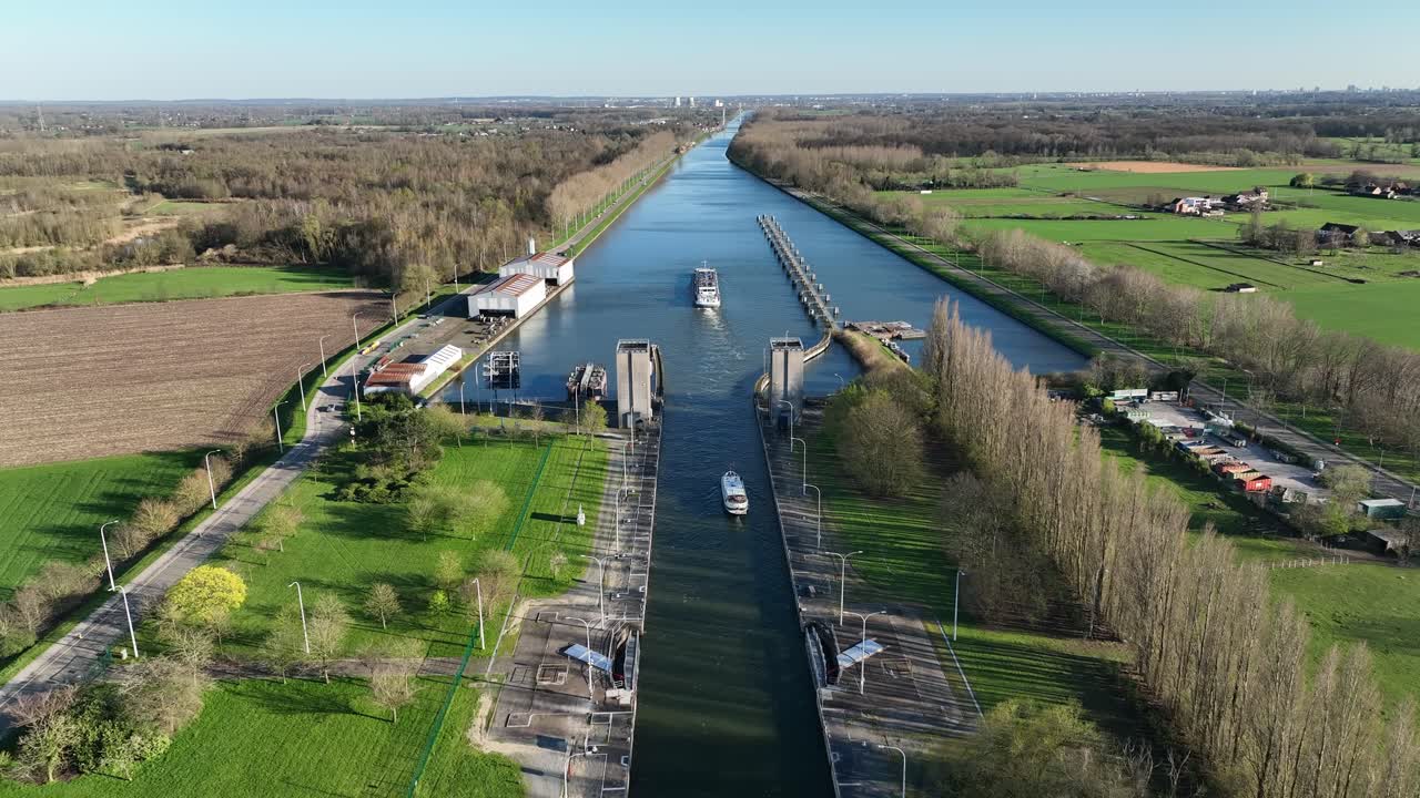 Zoom in aerial of Antwerp canal lock gate with boats crossing through waterways surrounded by green countryside and trees on a sunny day