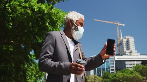 Hombre-Mayor-Afroamericano-Con-Mascarilla-Sosteniendo-Un-Teléfono-Inteligente-Y-Una-Taza-De-Café-Llamando-A-Un-Taxi-En-El