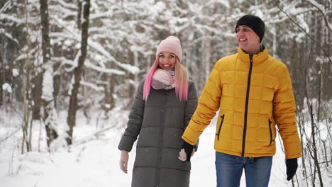 a man in a yellow jacket and a girl in a hat and scarf walk through the winter forest during a snowfall laughing and smiling at each other at christmas in slow motion