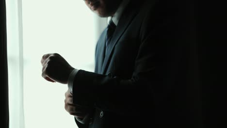 confident businessman in classic blue suit buttoning or adjust cufflinks near window in hotel room at the morning. handsome man wearing a nice suit on wedding day.