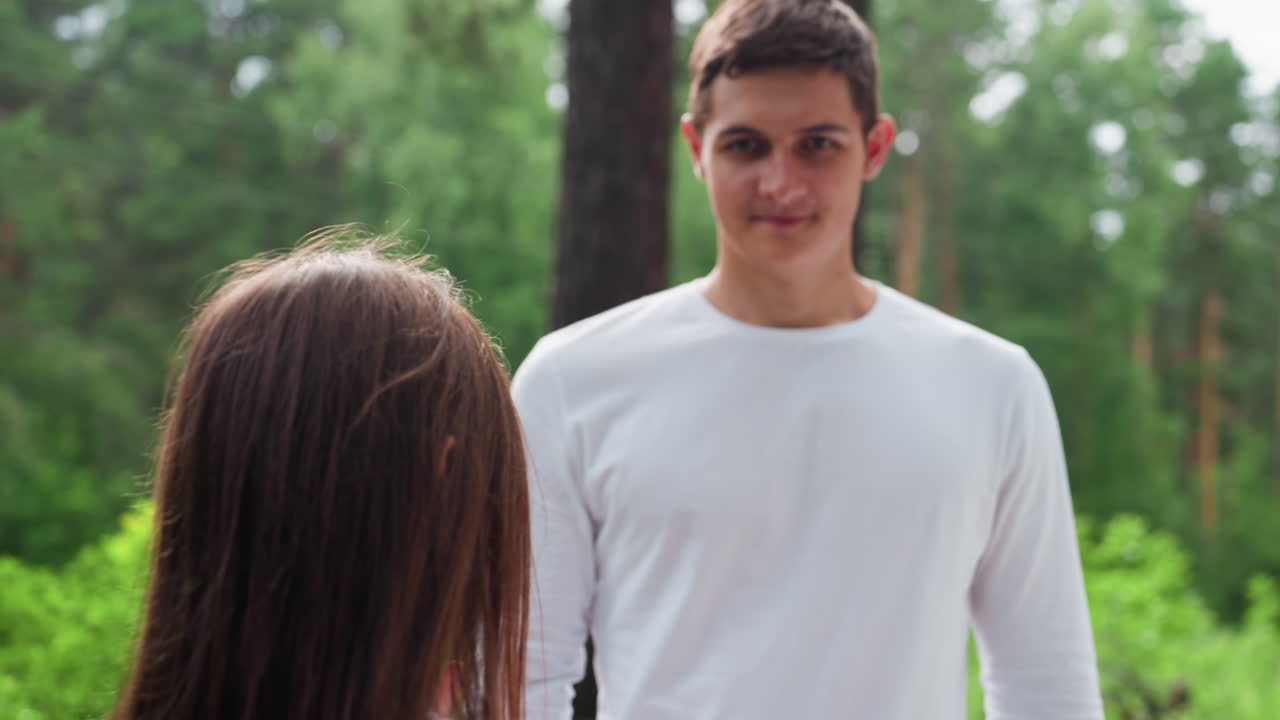 Una niña pequeña le entrega una cesta de picnic a su hermano mayor, que está de pie cerca de la zona de aparcamiento del bosque, rodeado de árboles verdes. Es una tranquila mañana de verano y la familia se prepara para un picnic, mostrando un gesto cariñoso al aire libre.