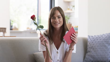 middle-aged caucasian woman smiles holding a rose and card at home on video call