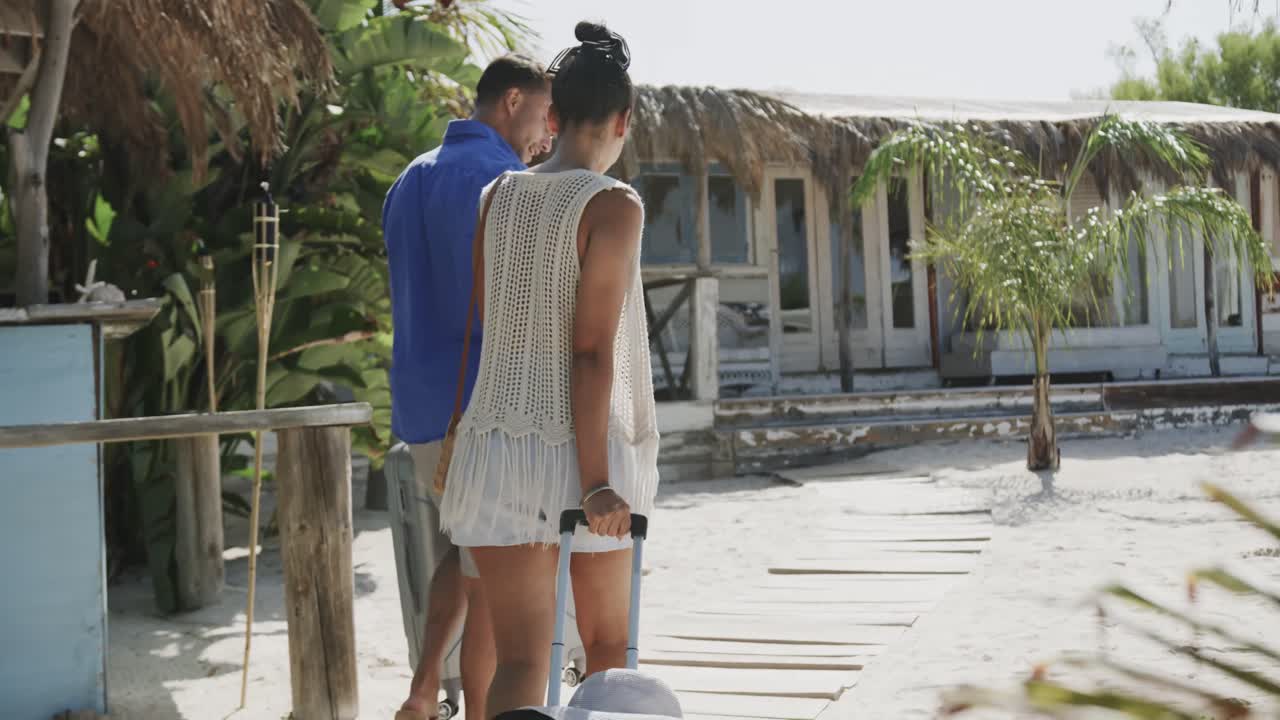 vista trasera de una feliz pareja diversa hablando, caminando con maletas en una playa soleada, en cámara lenta
