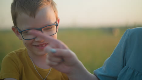 a young boy dressed in yellow playfully distracts his partially visible mother, who is sitting beside him in a grassy field. the boy smiles mischievously, enjoying the interaction with his mom