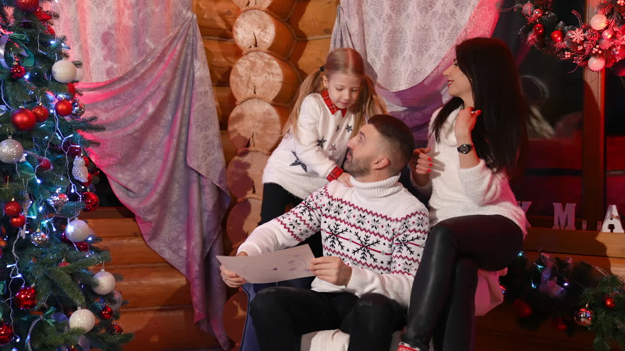 Family at Christmas night. Cheerful parents and their daughter spending time together in cozy living room. Little girl sits on father's shoulders. Happy family moments at winter holidays.