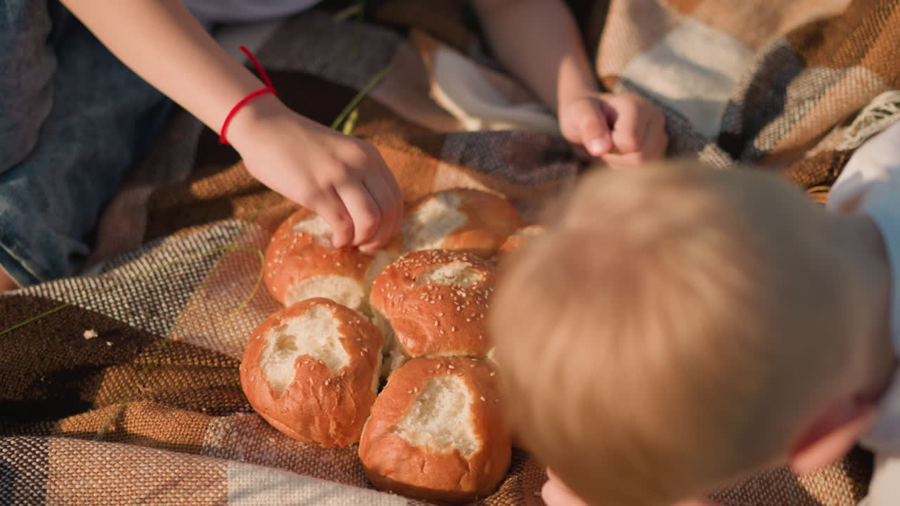 Close-up view of two young boys, faces not visible,pinching and eating bread from a cluster of rolls on a checkered picnic blanket. The scene captures a quiet, intimate moment of sharing food outdoors