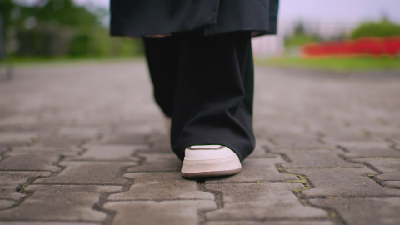 Close up front view of a female leg walking on stone pavement in black pants and white sneakers, low angle view of steps moving forward on cloudy rainy day with blurred greenery in background