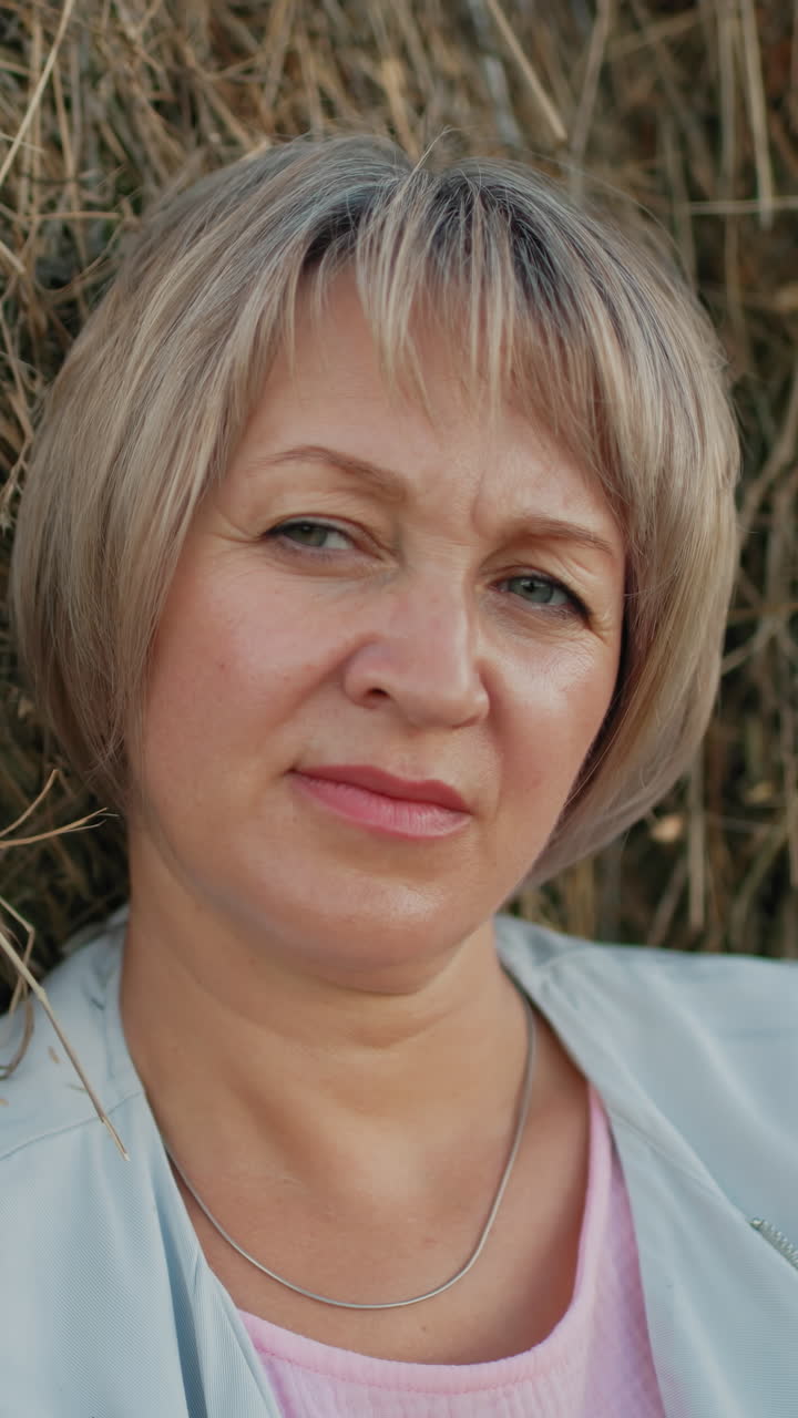 una mujer de mediana edad descansa pensativa en una bala de heno en una vasta tierra de cultivo, mira a la distancia, rodeada de campos ondulados, creando un momento rural pacífico y reflexivo bajo un cielo abierto.