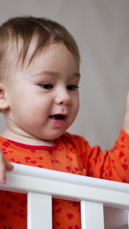 beautiful caucasian baby boy stays up in white cot. cute kid playing with pacifier throwing it on the floor and watching it fall. low angle view. vertical video