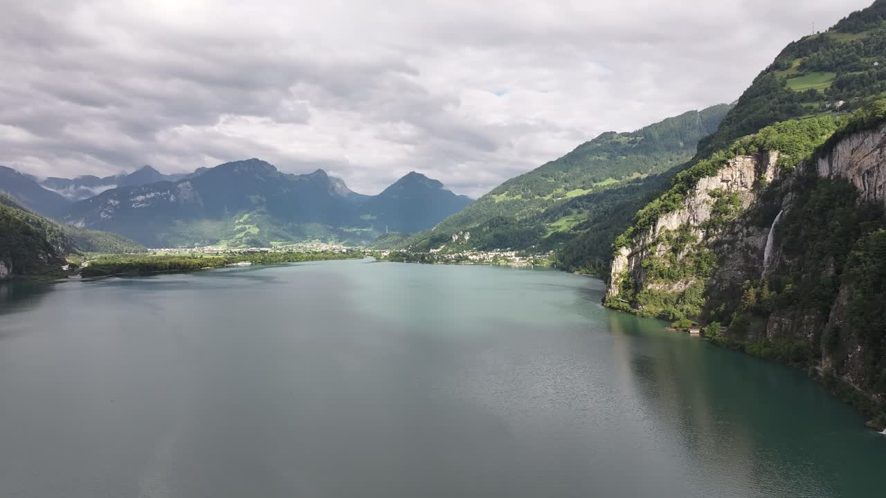 Scenic aerial view of Walensee lake in Switzerland, with towering cliffs, cascading waterfall, and lush alpine mountains under dramatic clouds