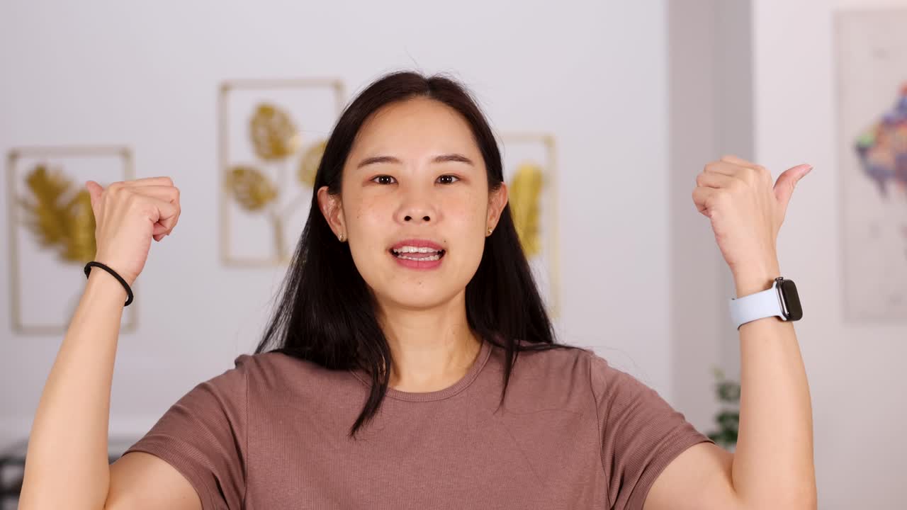 Young Asian woman smiles and gives double thumbs up in a bright, modern room with soft lighting and steady camera framing, conveying positivity
