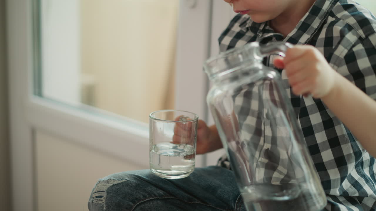 Close up of little boy wearing plaid shirt holding glass jar with left hand and glass cup with right hand carefully pouring water into cup while sitting by window indoors in bright daylight