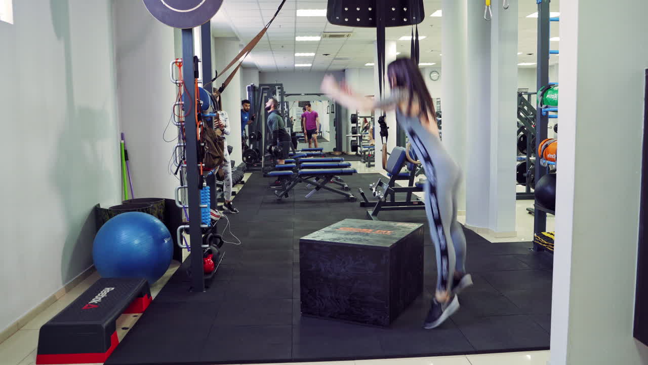 Athletic woman does jumps on a big box on the background of the modern fitness gym. Intense exercise jumping of a young female as her daily cross fitness training program.