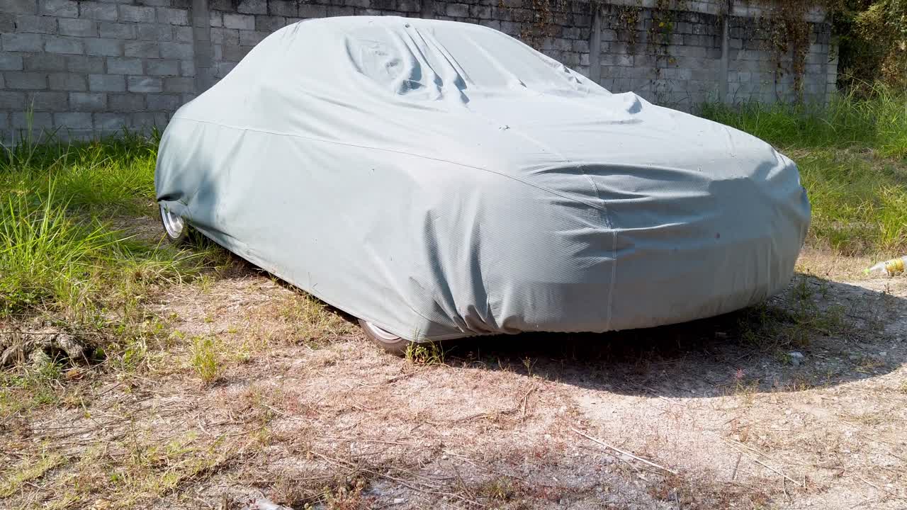 Un véhicule complètement recouvert d’une housse de voiture grise garé à l’extérieur sur de l’herbe près d’un mur de briques