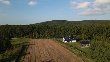 aerial backwards shot of farmland field with luxury house and solar panels on roof in rural suburb of poland - beautiful green forest trees in background