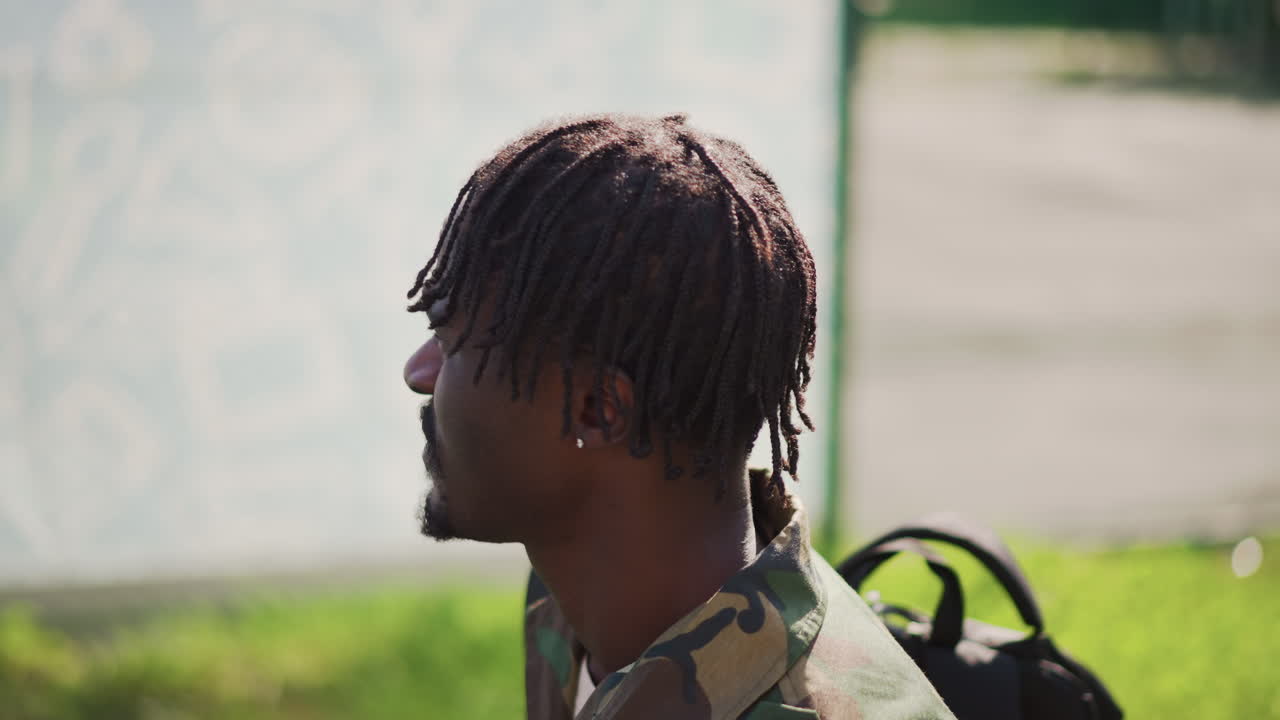 Reflective Soldier Walking, Veteran With Backpack Beside Fence, Quiet Moment Captured Of Military Veteran Reflecting, Contemplative Veteran In Camouflage Gear Walks Past Fence During Evening Hours