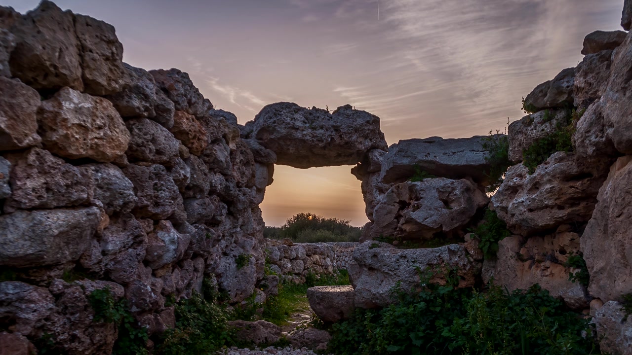 Il sole tramonta centrato nell'arco roccioso di una finestra delle Rovine di Trepuco a Minorca, Spagna, la luce nel cielo sfuma nell'ora blu