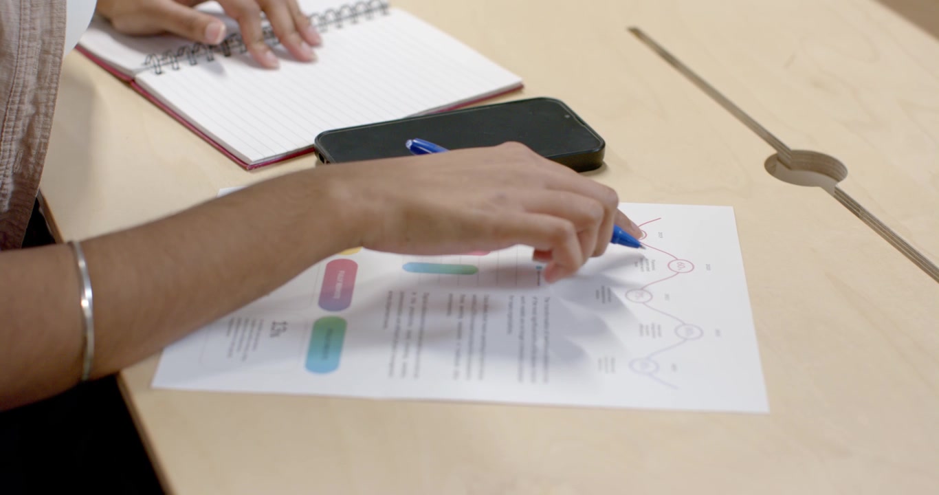 Indian woman opening notebook and tracing infographic with blue pen at table, reviewing for meeting