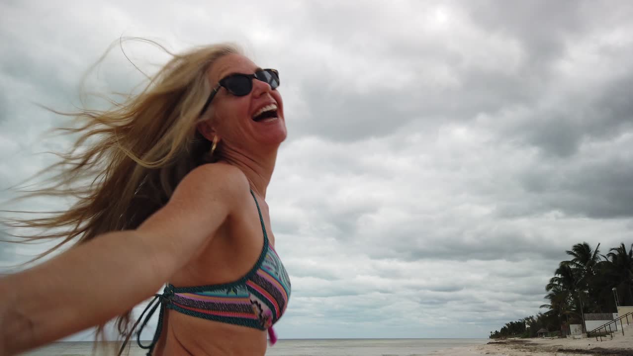 성숙한 여인은 해변에서 남편을 끌어당기는 나를 따라 슬로우 모션으로 비키니와 선글라스를 달린다