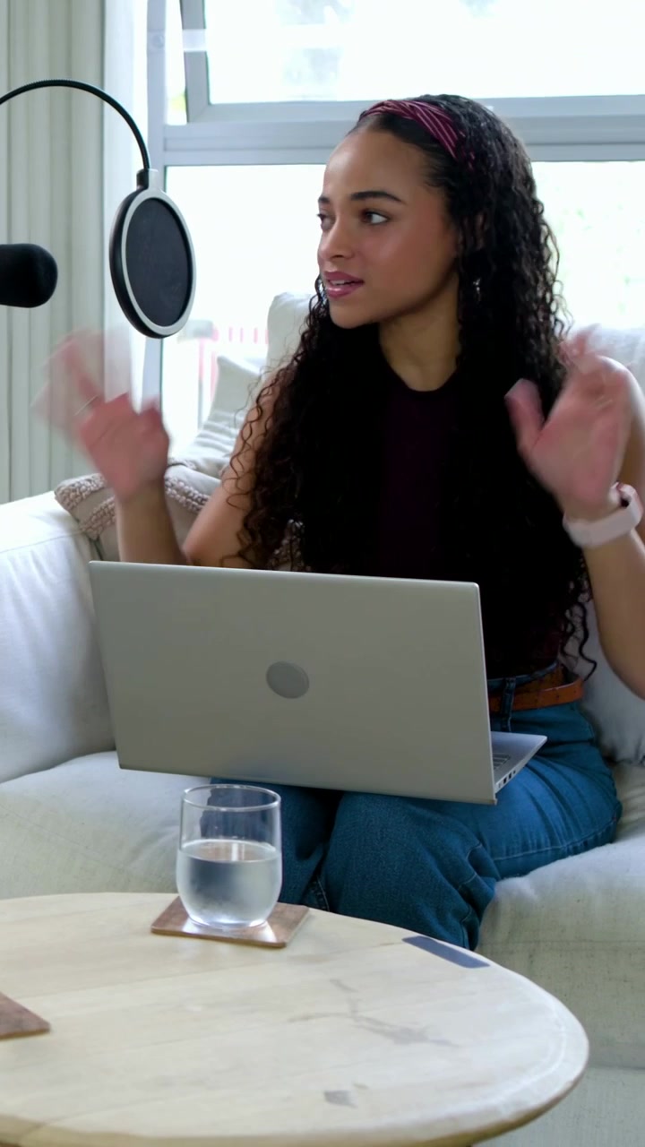 Video vertical: Mujer en camiseta de tirantes escribiendo, mirando el micrófono, hablando para un podcast en su portátil en casa.