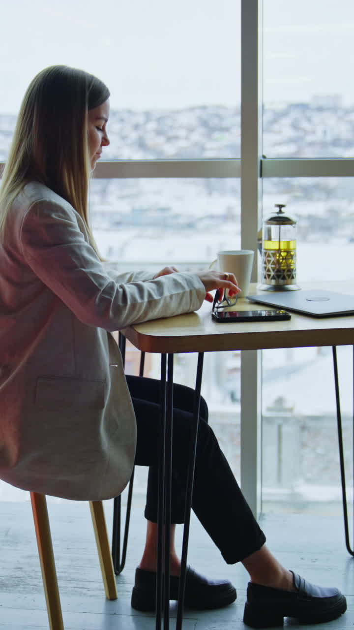 Confident happy woman sitting at table and having tea. Beautiful woman enjoying lunch break time at work. Side view. Vertical video