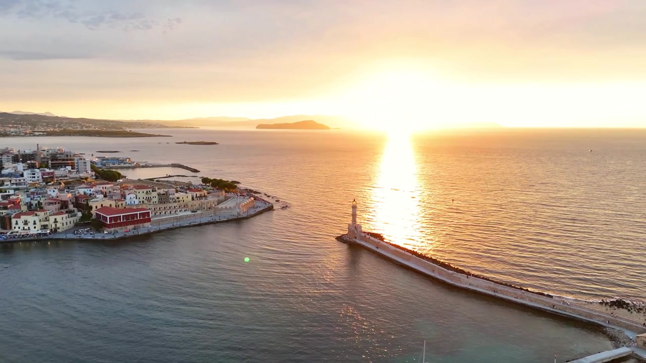 La telecamera aerea sorvola il frangiflutti di Chania al tramonto, con il faro, l'ingresso del porto, il lungomare della città vecchia e un brillante riflesso dorato sul mare.