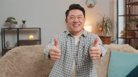 asian man sitting on sofa at home smiling and showing thumbs up feeling happy and satisfied