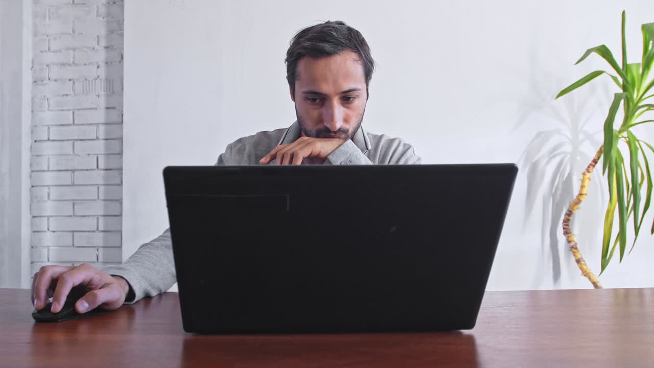 un hombre trabaja en una computadora portátil contra una pared blanca y una planta verde