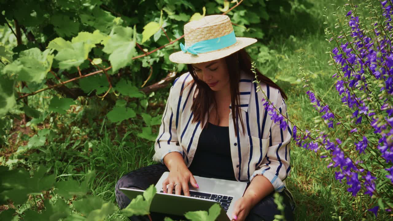 Donna con cappello di paglia che lavora al laptop tra fiori vivaci e una vegetazione lussureggiante in un ambiente sereno.