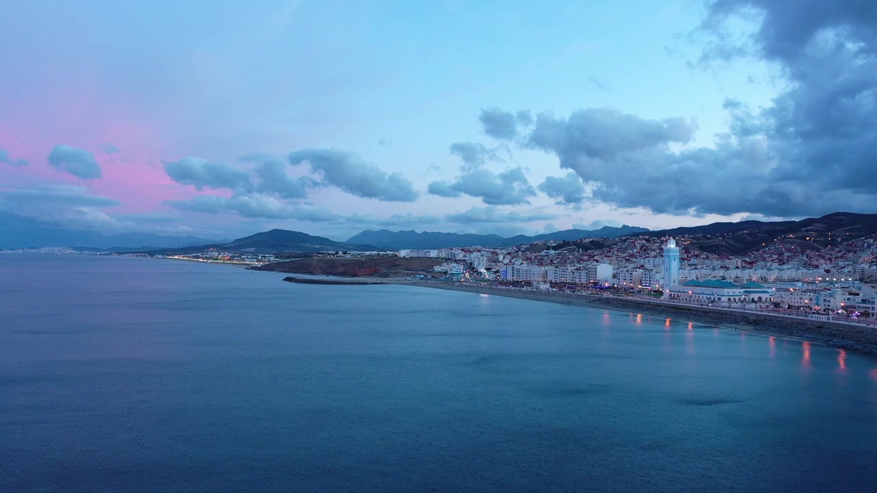 Ampia vista crepuscolare con drone sul Mar Mediterraneo verso M'diq, che mostra la costa illuminata, la moschea sul lungomare, i Monti del Rif e il cielo serale rosa nel Marocco settentrionale.