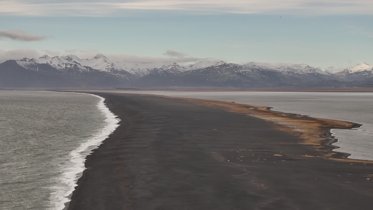 Aerial view of the long black sand coastline of Stokksnes Peninsula, Iceland, with waves crashing on one side and mountains rising in the background under a clear arctic sky.