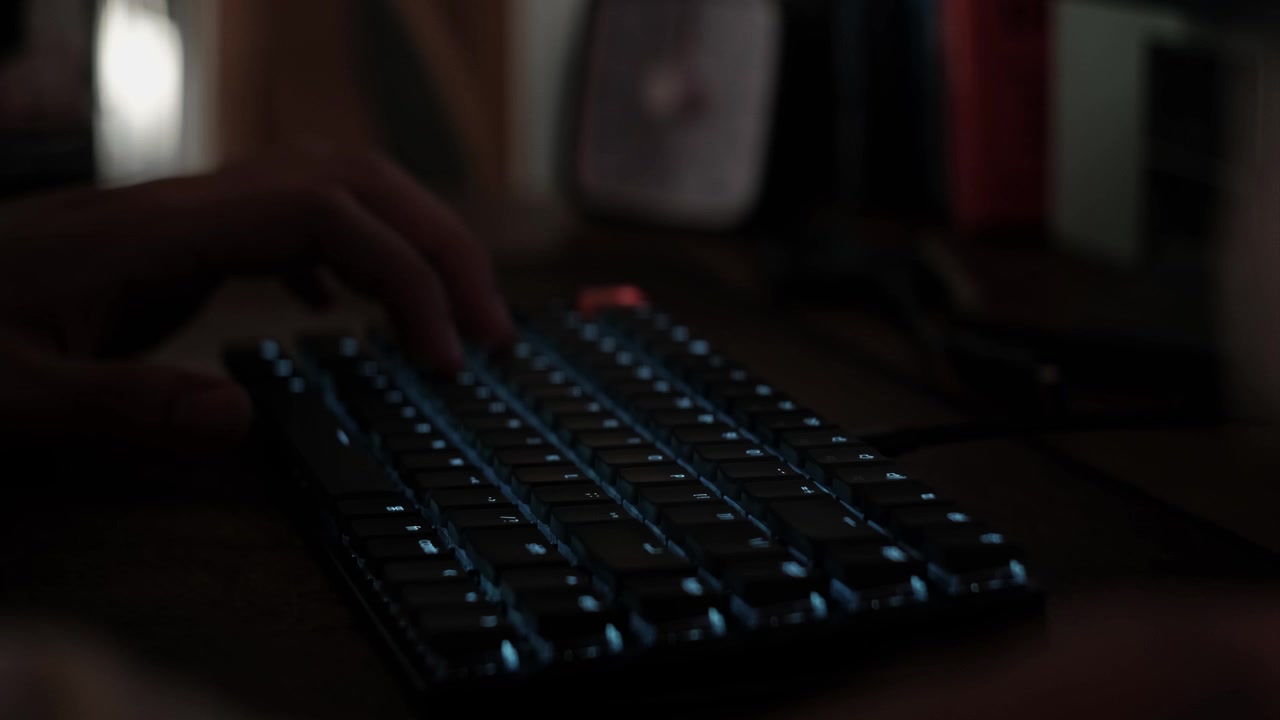 Close-up static shot of hands typing on a mechanical keyboard in a dim indoor workspace with illuminated keys and desk setup