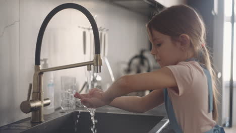 lavando las manos en la cocina niña pequeña en delantal está frotando sus palmas bajo el arroyo de agua del grifo