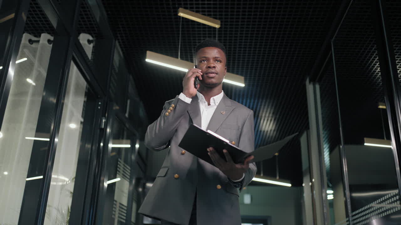 A black man is talking on the phone and walking down the corridor with documents