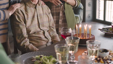 senior man blowing out candles on birthday cake during a celebration with his family at home while adult daughter filming a video with mobile phone