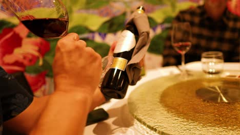 a person pours red wine into a glass at a gold coast dinner table, featuring warm lighting and a vibrant backdrop
