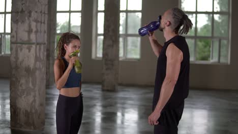 Stylish-sporty-man-and-woman-standing-in-studio-sport-club-with-windows-background-and-talking.-They-are-drinking-water-after-workout-with-color-bottles.-Both-with-stylish-dreadlocks