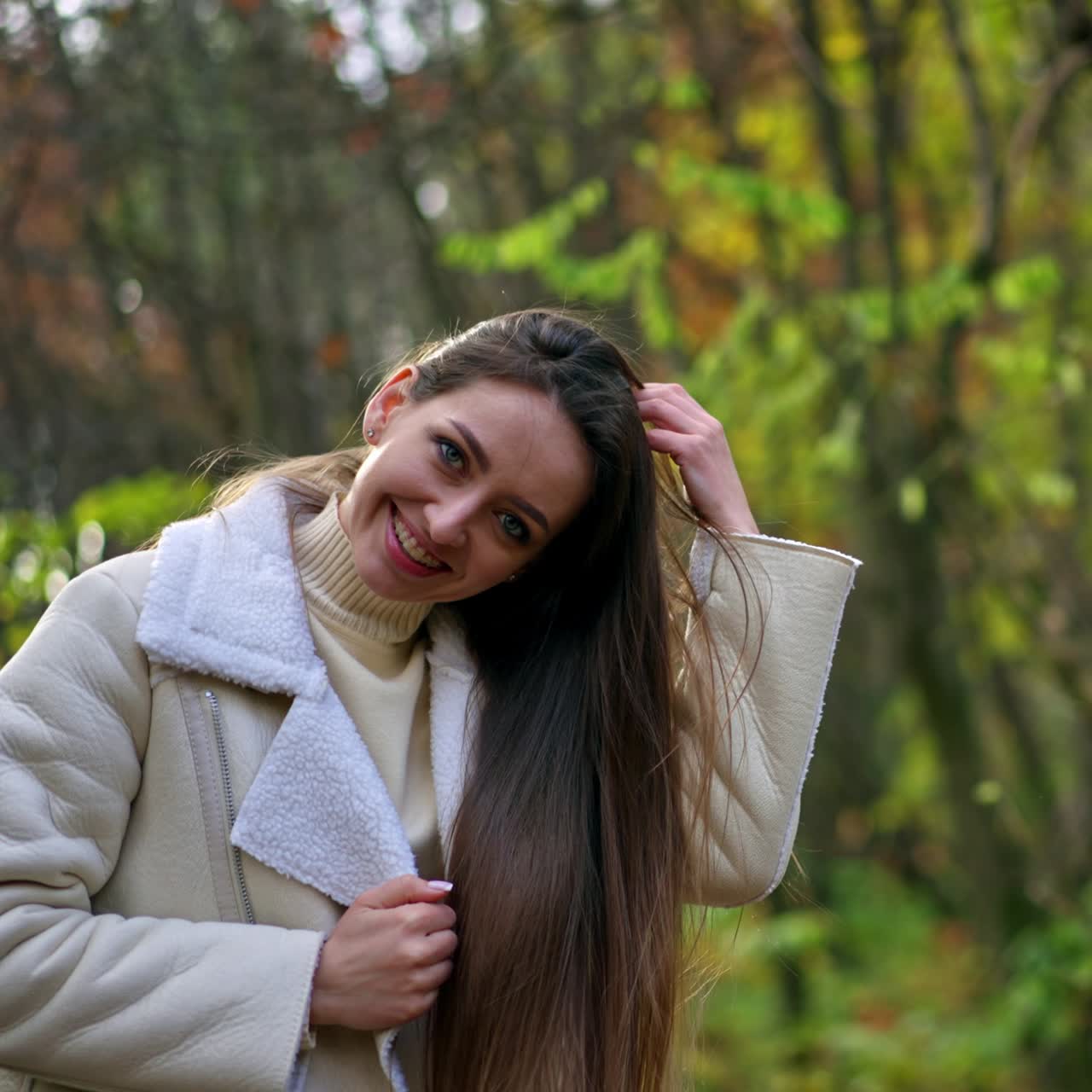 Smiling Caucasian lady in white jacket waving her long hair. Woman having fun relaxing in the walk at autumn park. Blurred backdrop