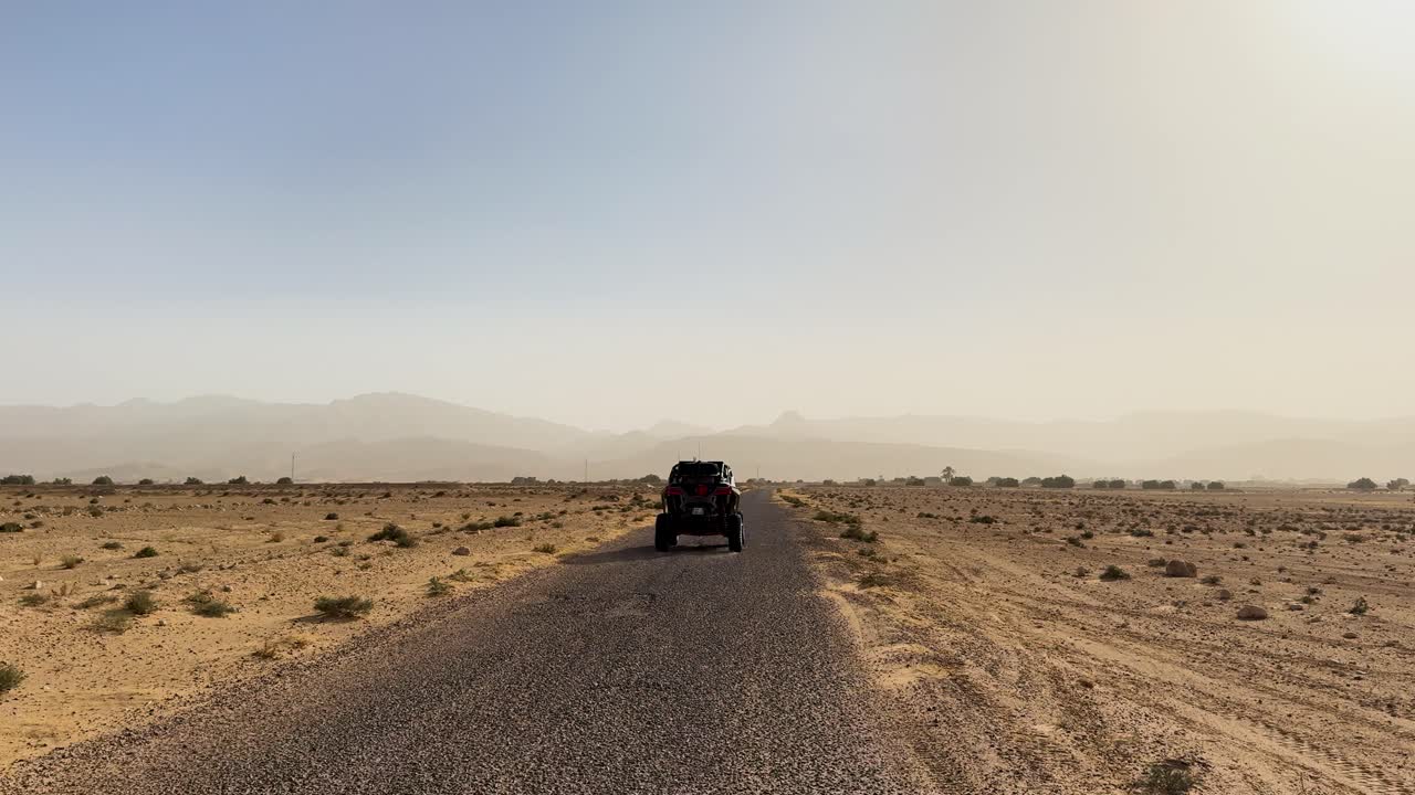 Vista posteriore di un fuoristrada che percorre una strada remota nel deserto con polvere di terreno arido, foschia, spazio aperto e montagne lontane sotto un cielo sereno diurno