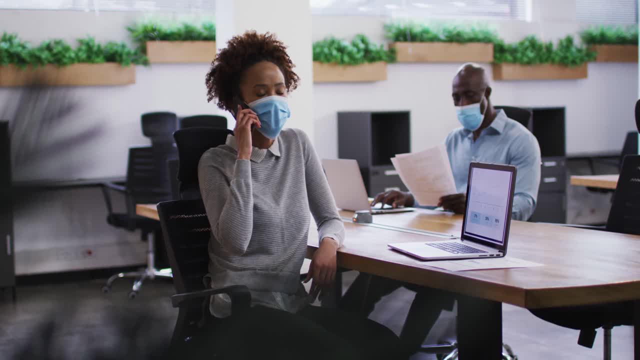 Diverse male and female business colleagues in face masks, woman talking on smartphone in office