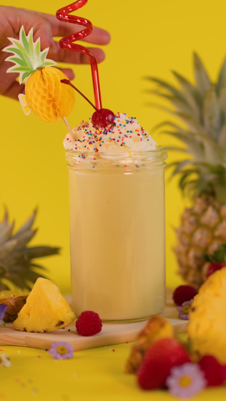 Una mano aggiunge una cannuccia a spirale decorativa a un frullato cremoso all'ananas, circondato da frutta, su sfondo giallo.