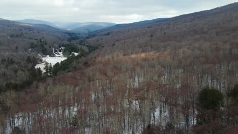 Imágenes-Aéreas-De-Un-Valle-De-Montaña-Invernal-Con-Nieve-Y-Dosel-Desnudo-Y-Montañas-Azules-En-La-Distancia