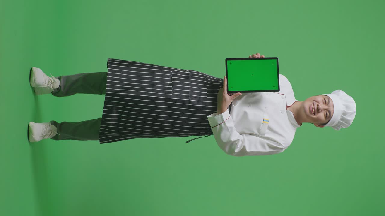 cuerpo lleno de hombre asiático chef sonriendo y mostrando la tableta de pantalla verde a la cámara mientras estaba de pie en el estudio de fondo de pantalla verde