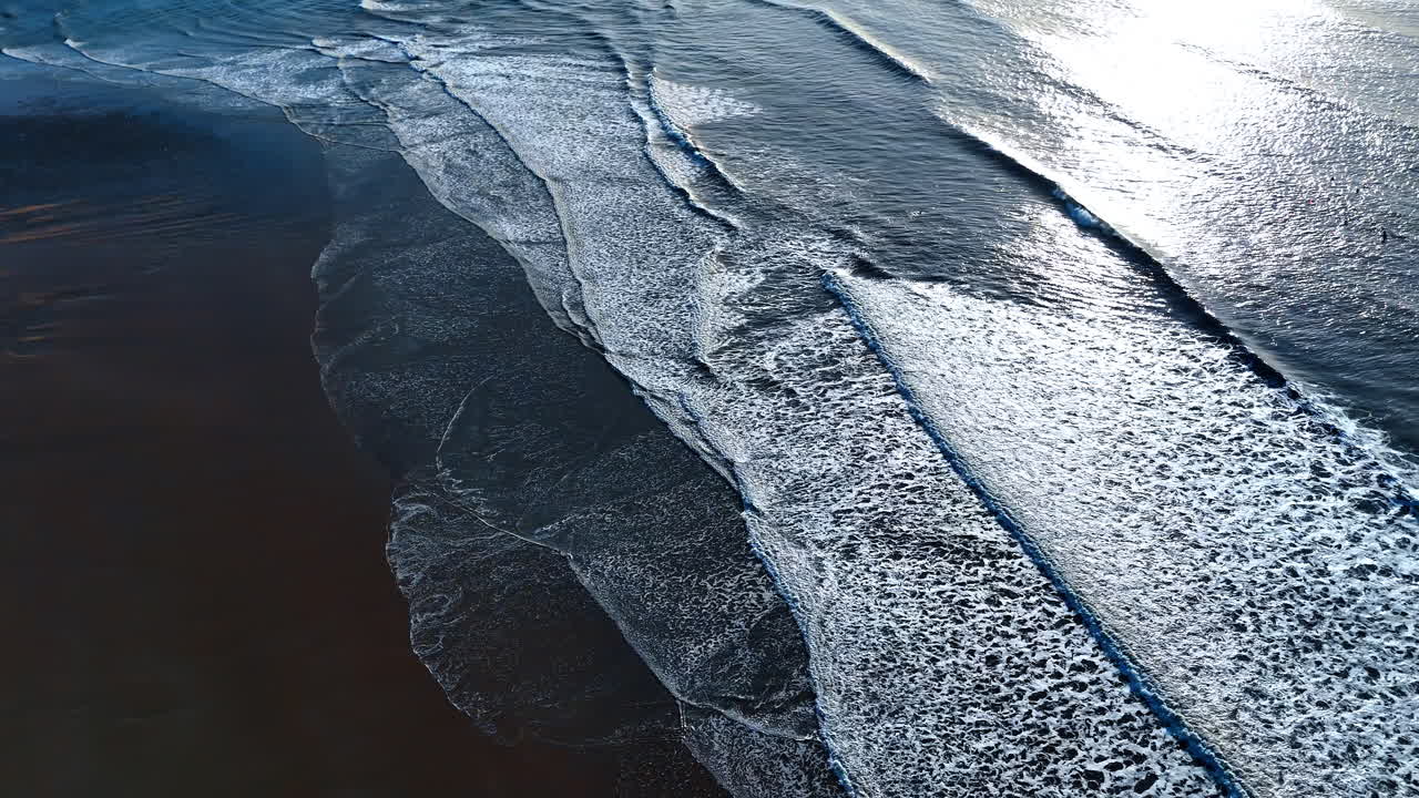 Aerial top down view of ocean waves on dark sand. Overhead perspective of white sea foam and breaking waves on a dark sandy beach during daylight