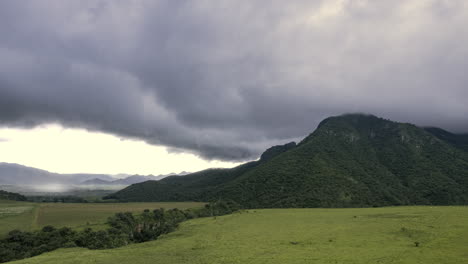 amplio campo verde con una imponente colina, luz solar en contraste y una tormenta a la izquierda, nubes de rápido movimiento que chocan con y rodean la colina, creando una sensación de velocidad y movimiento