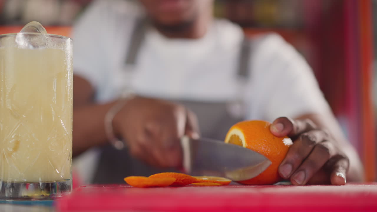 Barman cuts juicy orange on cutting board closeup. African American bartender prepares cocktail with citrus fruit for client. Fresh beverage preparation