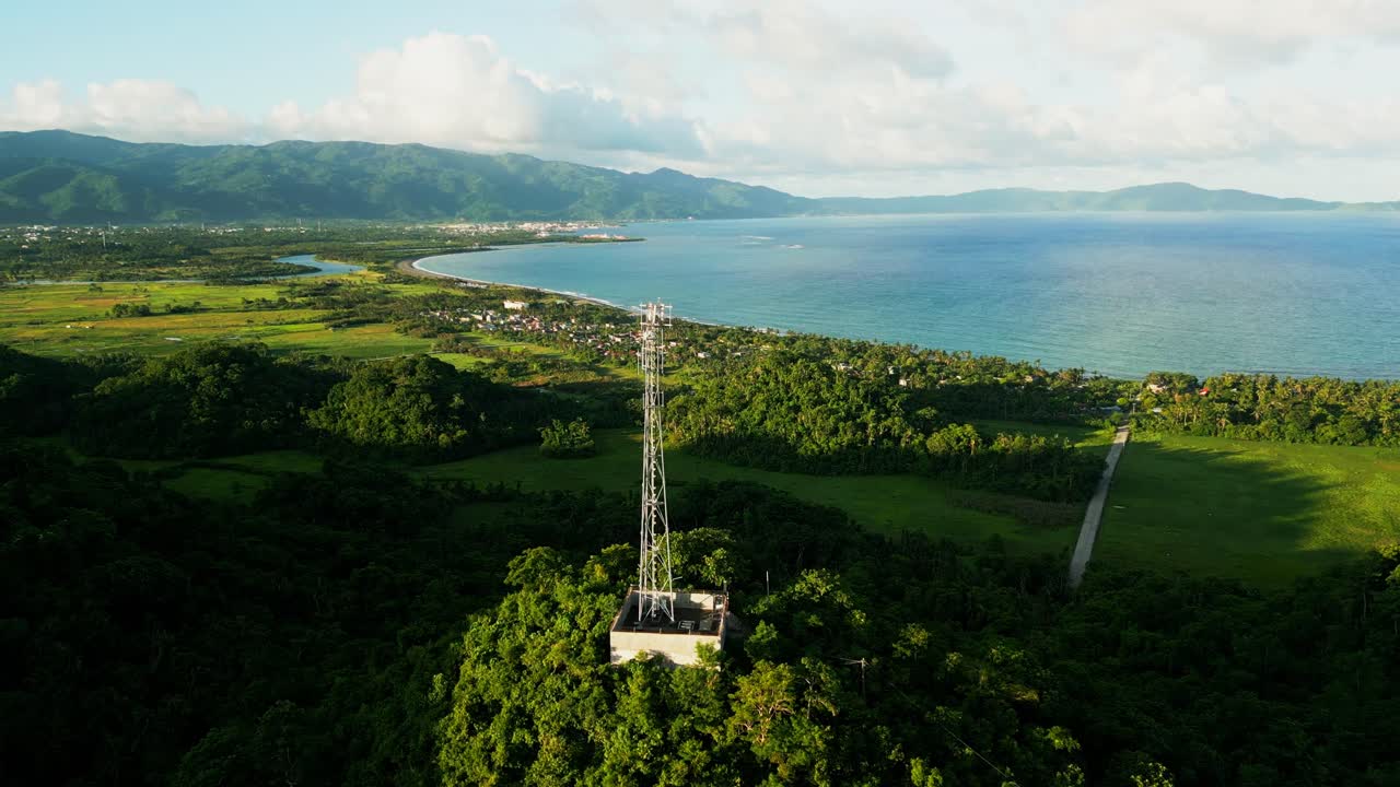 Scenic aerial drone shot of telecommunications tower atop lush hill overlooking tropical island village coastline at Virac, Catanduanes, Philippines.