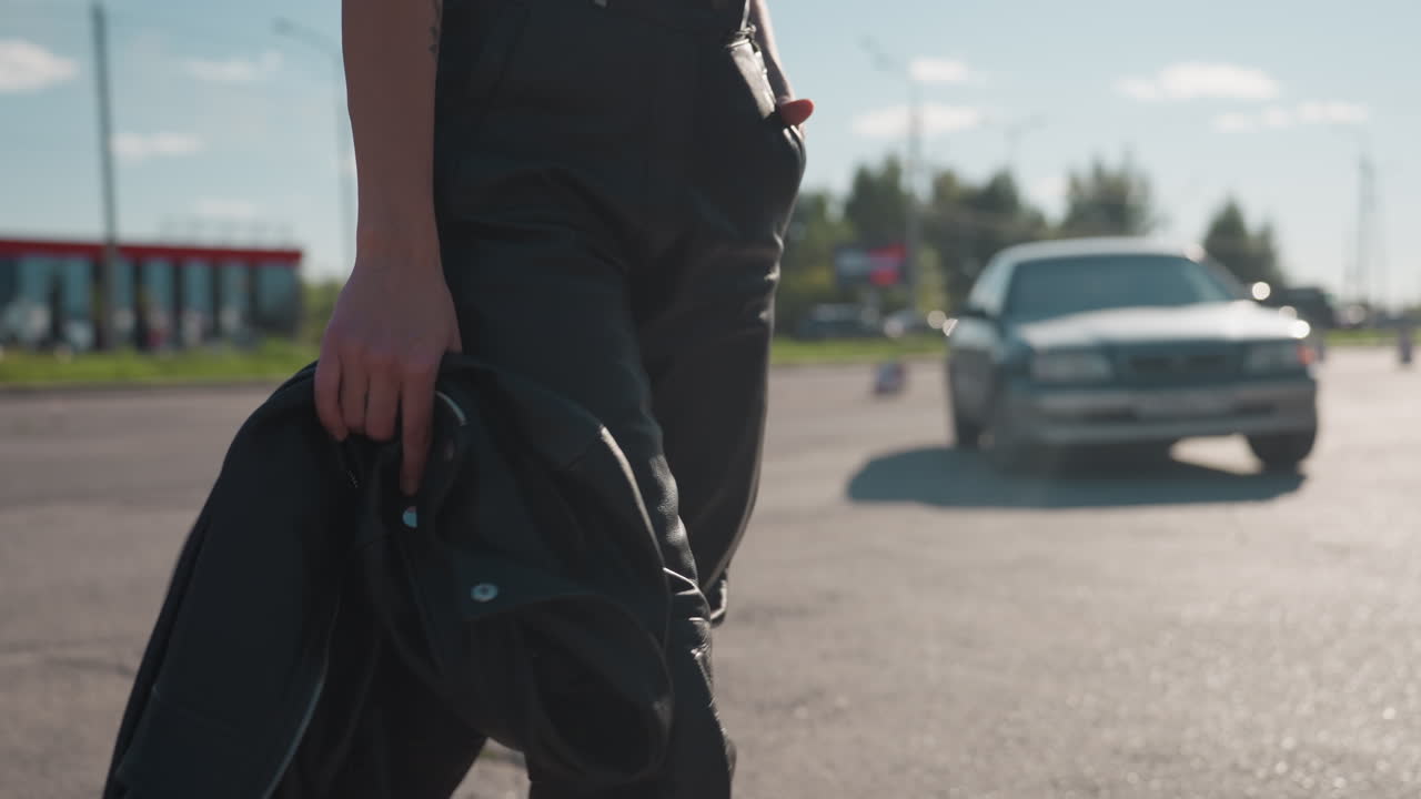 Close up side view of woman in black leather pants holding jacket while walking under sunlight with blurred cars and building in background, showing modern fashion and independence
