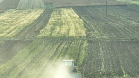 drone shot behind working tractor plowing green agricultural field during sunlight - animals following vehicle